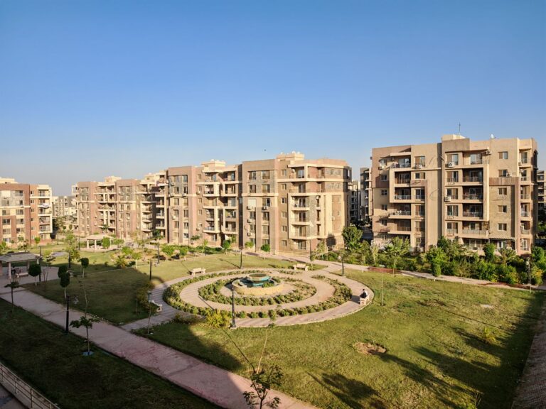 Mavi gökyüzü altında, ortasında peyzajlı bahçe ve yürüyüş yolları bulunan modern konut sitesi